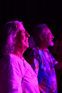 Two older adults stand side by side, smiling, on a dim stage bathed in purple light, watching a performance.