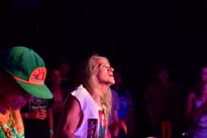 Musician with long blond hair and a white sleeveless shirt looks upward on stage under purple lighting, with a crowd in the dark background.