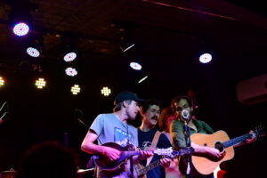 Three musicians on a dim stage: two with guitars and a vocalist at a vintage microphone under colorful stage lights.