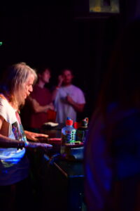DJ with long gray hair spinning at a dimly lit booth, colorful spray bottles and equipment on the counter, crowd in the background fades to shadowed figures in a club setting.
