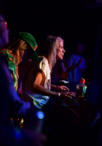 Older man with long gray hair leaning toward a keyboard on a dark stage, bathed in colorful lights, performing at a concert.