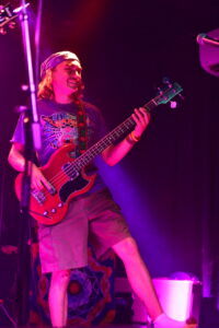 Guitarist wearing a bandana and t-shirt plays a red bass guitar on a dimly lit stage with pink-purple lighting.