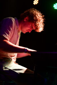 Young man playing a keyboard on a dimly lit stage with colorful lights overhead.
