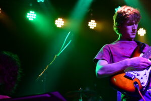 Male guitarist on a brightly lit stage playing an orange electric guitar, colorful green and purple lighting surrounding him.