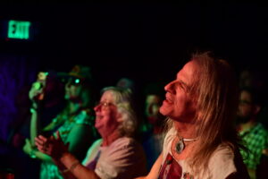 Front-row concertgoer with long blond hair smiling under colorful stage lights.