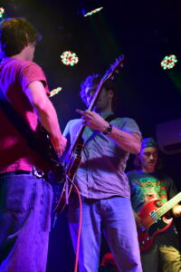 Two guitarists performing on a dimly lit stage; one in a pink shirt facing away, the other in a striped shirt playing an electric guitar under multicolored stage lights, with a bassist in the background.