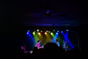 Band performing on a dimly lit stage with vibrant blue, green, and yellow lights, and a crowd of silhouettes in the foreground.
