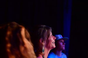 Profile of a smiling woman in colorful stage lighting, wearing long dangling earrings, with a blurred person in the foreground at a dark venue (likely a live event).