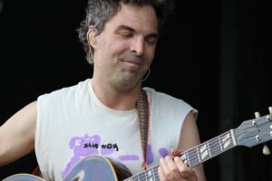 Male guitarist on stage with closed eyes, wearing a sleeveless shirt and colorful strap, playing an electric/acoustic guitar against a dark background.