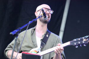 Guitarist with a shaved head and glasses singing into a microphone while playing an electric guitar on stage.