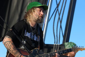 Guitarist with long blonde hair and a green cap playing an electric guitar on an outdoor stage against a blue sky, tattoos visible on arms.