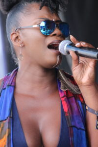 Close-up of a woman singing into a handheld microphone, wearing blue mirrored sunglasses and a colorful scarf.