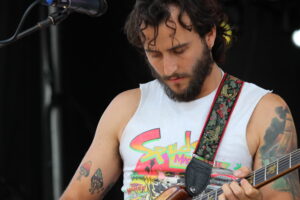 Male guitarist on stage, focused on playing a colorful guitar with a patterned strap and a white sleeveless shirt.