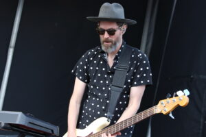 Male guitarist wearing a gray fedora, sunglasses, and a black polka-dot shirt, playing a cream-colored bass on stage with a keyboard nearby