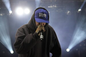 Performer in a dark hoodie wearing a blue cap reading 'Dirty', holding a microphone on stage with bright backlights behind him.