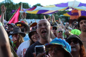 Crowd at an outdoor festival with colorful canopies overhead, people raising their hands and cheering toward the camera.