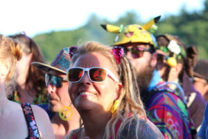 Smiling woman in white sunglasses in a colorful outdoor festival crowd, with vibrant outfits and accessories around her.