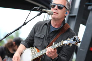 Male musician wearing sunglasses sings into a microphone while playing a sunburst resonator guitar on stage.