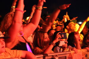 Audience at a nighttime concert with hands raised in the air and wristbands, one person filming with a smartphone under orange stage lighting.