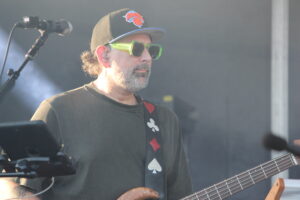 Musician wearing a Knicks cap and neon green sunglasses, playing bass on stage.