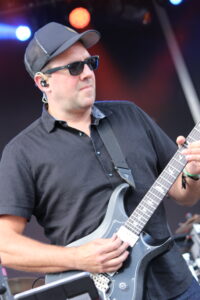 Male guitarist wearing sunglasses and a cap, playing a gray electric guitar on stage with colorful lights in the background