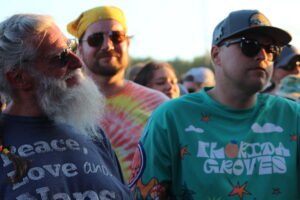 Group of people at an outdoor event, including an elderly man with a white beard on the left and two men wearing sunglasses in the foreground.