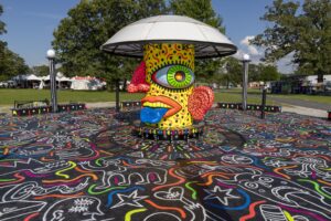 Colorful sculptural head with a dome hat in a park, surrounded by graffiti-patterned ground and festival tents in the background.
