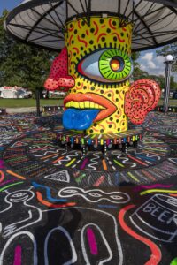 Colorful cartoonish face sculpture: yellow head with a large green eye, red nose, blue tongue, and pink polka-dotted ear on a patterned circular base in an outdoor setting.