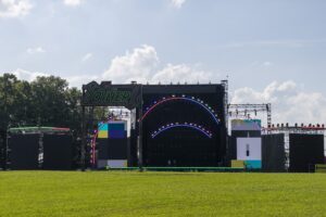 Outdoor concert stage in a grassy field with a neon-lit circular arch and a sign that reads 'The Other' under a blue sky.