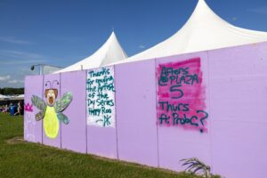 Purple graffiti wall with a cartoon bee; white event tents rise behind it under a bright blue sky.