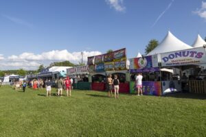 Outdoor festival scene with food stalls and white tents along a fence, people walking on grass under a blue sky edge of park campus fairground area.