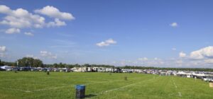 Open field at a festival or campground with rows of RVs, tents, and people along the horizon under a bright blue sky with fluffy clouds.