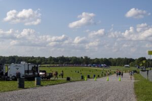 Open-field festival area with campers and tents along a gravel path, a parked RV on the left, and people walking under a bright, cloudy sky