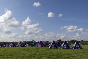 Row of colorful festival tents set up on a grassy field under a blue sky with scattered white clouds.
