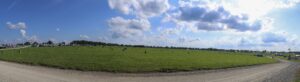 Panoramic view of a grassy field at an outdoor fair, with tents, vehicles, and a distant ferris wheel under a blue sky.