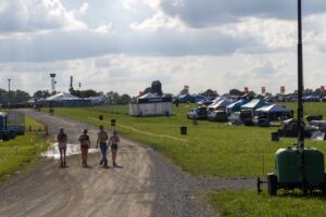 Group of four young people walking toward the camera along a muddy festival path with tents and parked cars in the background under a cloudy sky.