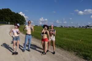 Four friends in swimsuits pose on a dirt road at an outdoor festival, with tents and a blue sky in the background.