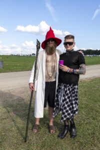 Two festival-goers pose together outdoors: one in a red wizard hat, white robe, and staff; the other in a black top, spiked sunglasses, and plaid skirt.