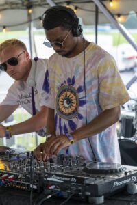 DJ wearing headphones and sunglasses mixers tracks on a Pioneer DJ setup at an outdoor event under string lights.