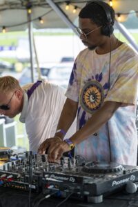 DJ wearing headphones and tie-dye shirt mixing on Pioneer DJ equipment at an outdoor event.