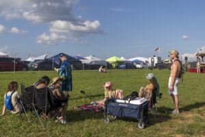 Group of festival-goers relaxing on a grassy field with colorful tents and parked cars behind a chain-link fence.
