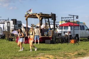 Outdoor festival scene with a makeshift DJ booth on a trailer and attendees in swimwear strolling on grass under a blue sky.