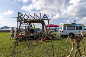 Outdoor festival with a large wooden geometric sculpture being assembled; tents and a white van in the background.] ,