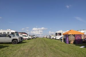 Row of white RVs parked on a grassy field at an outdoor RV event, with an orange patterned tent visible on the right.
