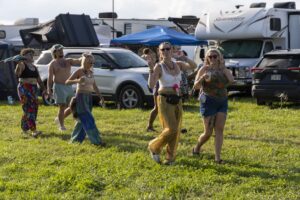 Group of festivalgoers dancing on a grassy field with RVs and tents in the background on a sunny day, wearing colorful outfits.