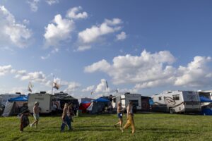 RV trailers and tents lined up on a grassy field under a blue sky with white clouds, colorful flags fluttering above. It’s a festival or gathering scene with people walking about and camp gear visible.