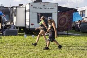 Two people in black outfits walk past a white RV labeled 'Trail Runner' at a grassy campsite with tents and banners nearby.