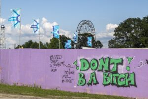 Purple graffiti wall in foreground with bold green lettering; flags, trees, and a Ferris wheel in the distance under a blue sky.