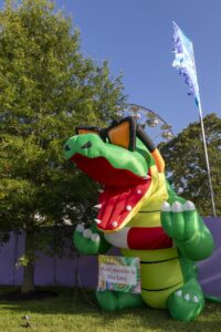 Large colorful inflatable dragon outdoors with a banner that reads 'Masturbation Is Self Love' and a Ferris wheel in the background.