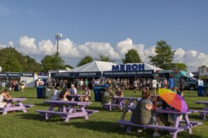 Crowd at an outdoor festival with a large white tent labeled MERCH and purple picnic tables on green grass under a blue sky.